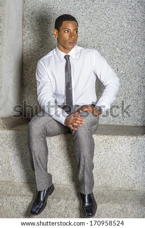 Dressing in white shirt, a black tie, gray pants, leather shoes, wearing a watch, hands resting on his lap, a young black businessman is sitting outside to take a break. / Relaxing Outside