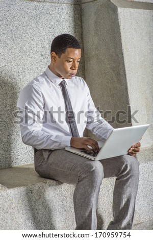 Dressing in white shirt, a black tie, gray pants, a young black college student is working on a computer outside an office building. / Working Outside