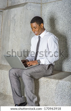 Dressing in white shirt, a black tie, gray pants, a young black college student is working on a computer outside an office building. / Working Outside