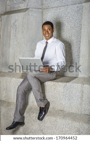 Dressing in white shirt, a black tie, gray pants, a young black college student is working on a computer outside an office building. / Study Outside