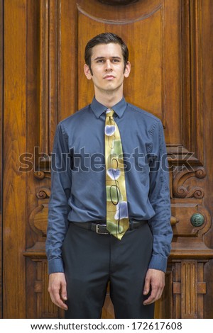 Dressing in a blue shirt, pants and a colorful tie, a young college student is standing in the front of old fashion style office door, looking forward. / Portrait of College Student