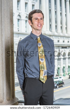 Dressing in a blue shirt, black pants, a colorful tie, hands putting in the back, a young college student is standing outside a business building, smilingly looking forward. / College Student