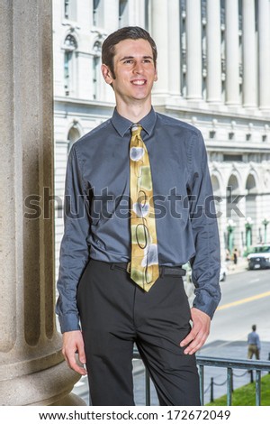 Dressing in a blue shirt, black pants, a colorful tie, a young college student is standing outside a business building, smilingly looking forward. / Portrait of College Student