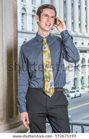 Dressing in a blue shirt, black pants, a colorful tie, a young college student is standing outside a business building, making a phone call. / Talking on Phone