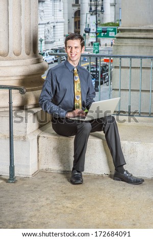 Dressing in a blue shirt, black pants, a colorful tie, a young college student is sitting outside on campus, smiling, working on a computer on his lap. / Study Outside