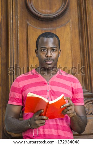 Wearing a red, pink stripe Henley V Neck shirt, gray pants, a bracelet, a young handsome black guy with a little mustache is reading a book by a old fashion style door. /Reading Book