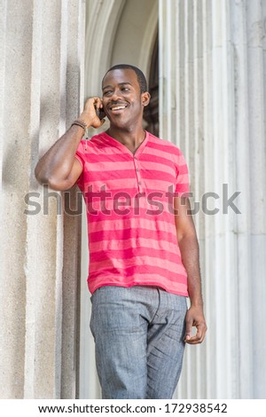 Dressing in red, pink stripe Henley V Neck T shirt,  gray pants, wearing a bracelet, a young black guy is standing by a column, smiling and making a phone call. / Calling Outside