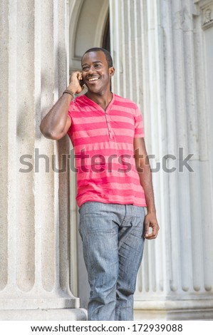 Dressing in red, pink stripe Henley V Neck T shirt,  gray pants, wearing a bracelet, a young black guy is standing by a column, smiling and talking on a cell phone. / Making Phone Call Outside