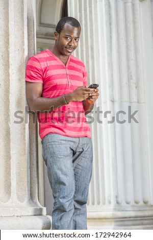 Dressing in red, pink stripe Henley V Neck T shirt,  gray pants, wearing a bracelet, a young black guy is standing by a column, checking messages on his smart phone. Text