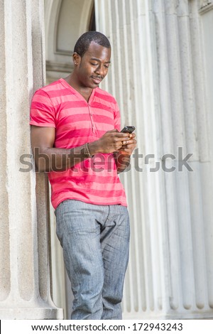 Dressing in red, pink stripe Henley V Neck T shirt,  gray pants, wearing a bracelet, a young black guy is standing by a column, checking messages on his smart phone. / Text