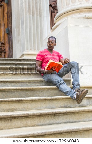 Stretching one leg, holding a red book, a young black guy is sitting on stairs outside an office building, reading. / Reading Outside