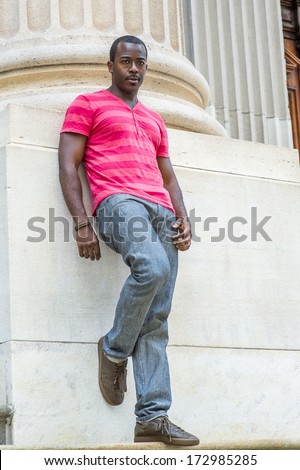 Wearing a red, pink stripe Henley V Neck T shirt,  gray pants, leather sneakers, bending over a leg, a young black guy is standing against the base of a column, relaxing and thinking.