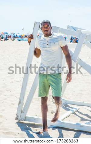 Wearing a white short sleeve Henley shirt, green shorts, barefoot, a young black guy is standing by a white wooden structure on the beach, thoughtfully looking at you. Many people on the background.