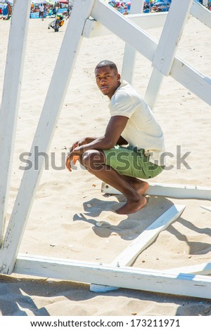 Wearing a white short sleeve shirt, green shorts, barefoot, a young handsome black guy is squatting under a white wooden structure on the beach, looking around, waiting for you.