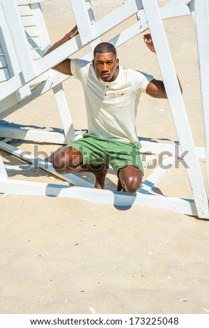 Wearing a white Henley shirt, green shorts, barefoot, a young handsome black guy is squatting under a white wooden structure on the beach, thoughtfully looking at you. / On the beach