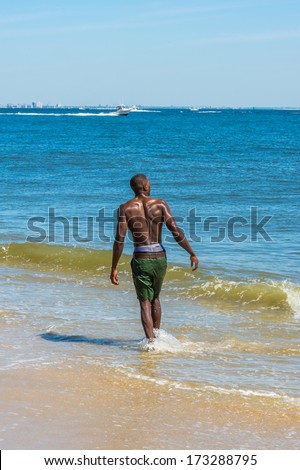 A young black guy, half naked, whole body wet,  is walking on water of ocean, looking forward. The horizontal is a big city outline, a small boat in the background. / Walking on the beach