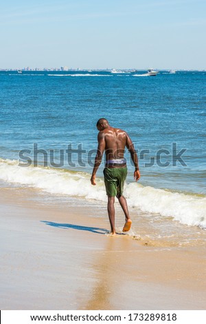 Looking down, half naked, whole body wet, a young black guy is walking on the beach. The horizontal is a big city outline, small boats on the background. / Walking on the beach