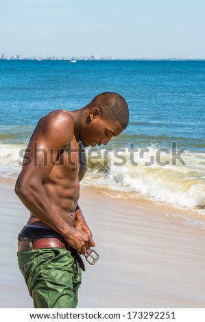 A well built young sexy black guy, half naked,  is taking off his wet pants on the beach. / Taking Off