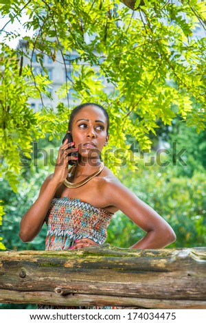 Dressing in a strapless colorful dress, wearing a golden neck ring, a pretty black woman is making a phone call inside small words. / Making Phone Call Outside