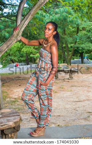 Dressing in a strapless fashionable, contemporary style dress top, pants, fashionable sandals, a hand putting in a pocket, a pretty black woman is standing by a tree in a park, relaxing. /Fashion Girl