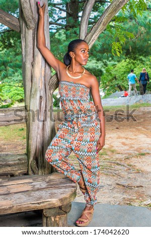 Dressing in a strapless fashionable, contemporary style dress top and pants, fashionable sandals, a pretty black woman is standing by old trees in a park, relaxing. / Relaxing in Park