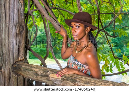 Wearing a brown outback hat,a golden neck ring,  a pretty black woman is standing outside in a small woods, looking at you. / Portrait of  Pretty Black Girl