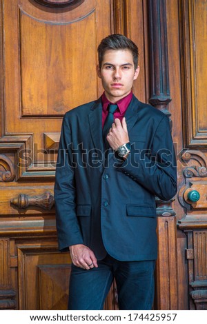 Dressing in a red undershirt, a black blazer, black jeans and a black tie, a young handsome college student is standing in the front of old fashion style door, looking forward. / Young College Student