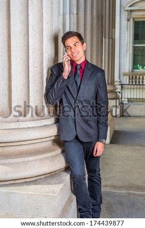 Dressing in a red undershirt, a black blazer,  jeans,  tie, a young handsome college student is leaning against a column outside an office, smiling, making a phone call. / Calling Outside
