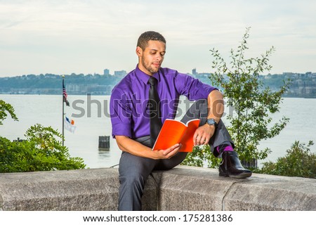 Dressing in a purple shirt, gray pants and a black tie, holding a red book, a young college student with a little beard and mustache is sitting by a river,  reading outside on campus. / Study Outside