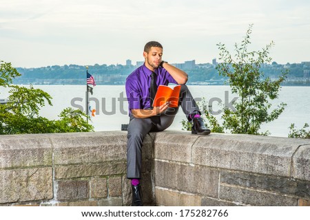 Dressing in a purple shirt, gray pants and a black tie, holding a red book, a young college student with a little beard and mustache is sitting by a river,  reading outside on campus. / Study Outside