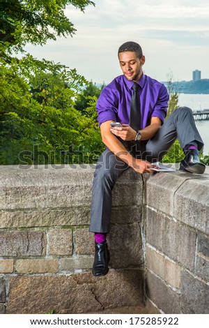 Dressing in a purple shirt, gray pants, a black tie, holding a red book, a college student with a little beard and mustache is sitting by a river on campus, checking messages on his smart phone./Text