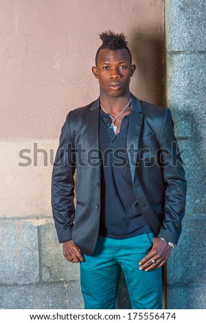 Dressing in a black blazer, green pants, a young black guy with mohawk hair is standing against the wall, confidently looking at you. / Portrait of Young Black Guy