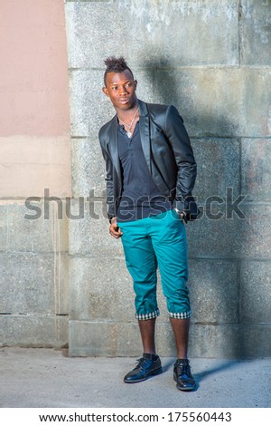 Dressing in a black blazer, green pants, leather shoes,  a hand in his pocket, a young black guy with mohawk hair is standing against the wall, looking forward. / Portrait of Young Black Guy