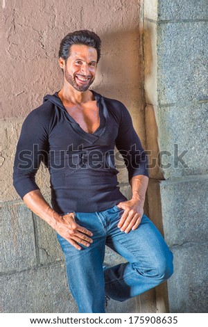 Dressing in a black sweater and blue jeans, a handsome, sexy, middle age guy with mustache and beard is leaning against the wall, smilingly looking at you. / Portrait of Middle Age Guy