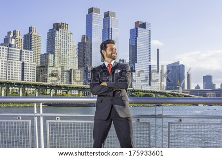 Dressing is a black suit, a red tie, crossing arms, a sexy, middle age businessman with mustache and beard is standing in the front of a business district, confidently looking forward. /Businessman