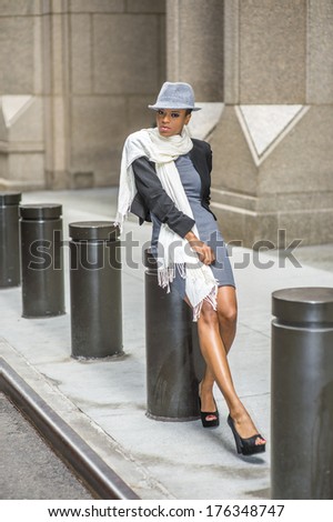 Dressing in a cropped jacket,  a fitted dress, open toes shoes and a long scarf, wearing a woolen Fedora hat, a young black woman is sitting on a metal column outside a building, relaxing.