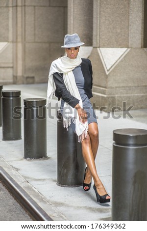 Dressing in a cropped jacket,  a fitted dress, open toes shoes and a long scarf, wearing a woolen Fedora hat, a young black woman is sitting on a metal column outside a building, relaxing.