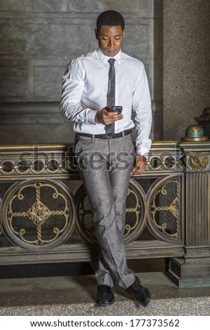 Wearing a white shirt, a black tie, gray pants, a young handsome black college student is standing by a railing on campus,  checking messages on a mobile phone. / Text