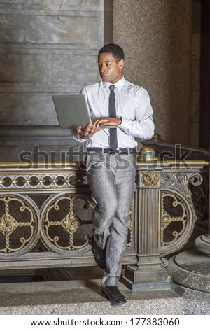 Wearing a white shirt, a black tie, gray pants, a young handsome black college student is standing by a railing in a hallway on a campus, working on a laptop computer. /Study Anywhere