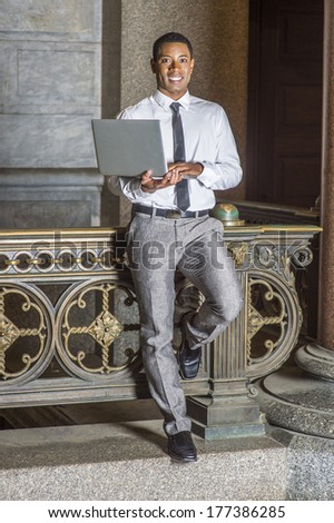 Wearing a white shirt, a black tie, gray pants, holding a laptop computer, a young handsome black college student is standing by a railing in a hallway on a campus, smiling, looking at you.