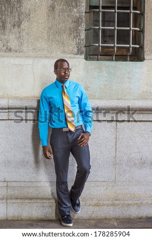 Dressing in a light blue shirt, a pattern tie, gray pants, wearing glasses, a black college student is standing by a pattern wall with a window, confidently looking forward. /College Student