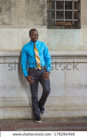 Dressing in a light blue shirt, a pattern tie, gray pants, wearing glasses, a black college student is standing by a pattern wall with a window, confidently looking forward. /College Student