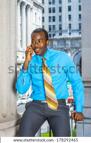 Dressing in a light blue shirt, gray pants,  a pattern tie, a young black businessman is sitting outside an office building, making a phone call. / Calling Outside