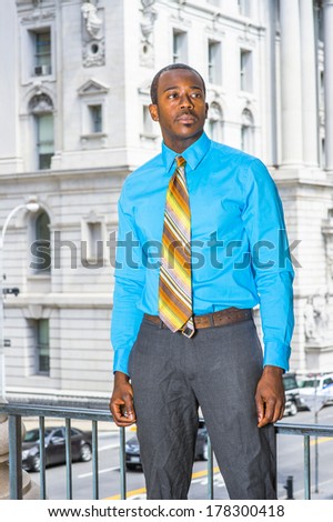Dressing in a light blue shirt, gray pants,  a colorful pattern tie, a young black businessman is standing outside an office building, looking forward. / Portrait of Young Businessman