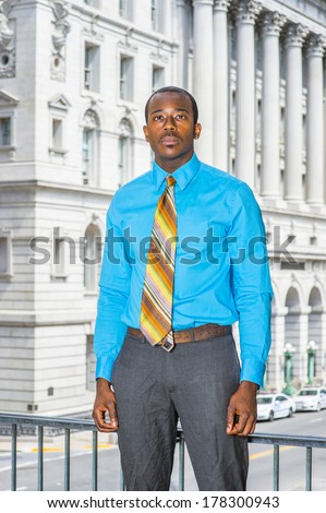 Dressing in a light blue shirt, gray pants,  a colorful pattern tie, a young black businessman is standing outside an office building, looking forward. / Portrait of Young Businessman