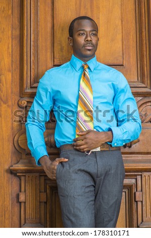 Dressing in a light blue shirt, gray pants,  a colorful pattern tie, a hand putting in pocket, a young black businessman is standing by a old fashion style door, lost in thought. /Black Businessman