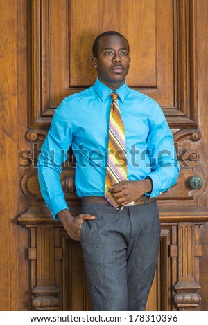 Dressing in a light blue shirt, gray pants,  a colorful pattern tie, a hand putting in pocket, a young black businessman is standing by an old fashion style door, lost in thought. /Black Businessman