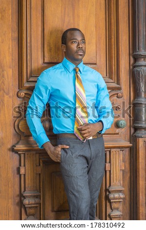 Dressing in a light blue shirt, gray pants,  a colorful pattern tie, a hand putting in pocket, a young black businessman is standing by a old fashion style door, lost in thought. / Black Businessman