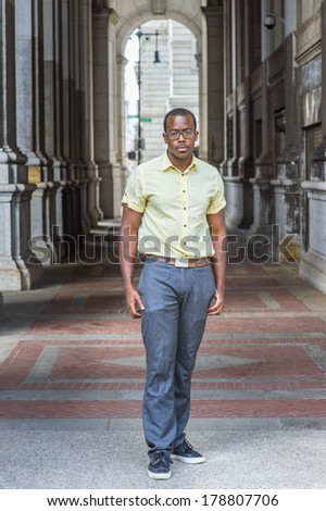 Dressing in a light blue shirt, a pattern tie, gray pants, wearing glasses, a black college student is standing on a walkway on campus, looking at you. / Portrait of College Student