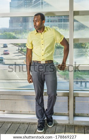 Wearing a light yellow shirt, gray pants and black shoes,  a young black guy is standing by a big glass wall into deeply thinking. The background is over a street scene. /City Life